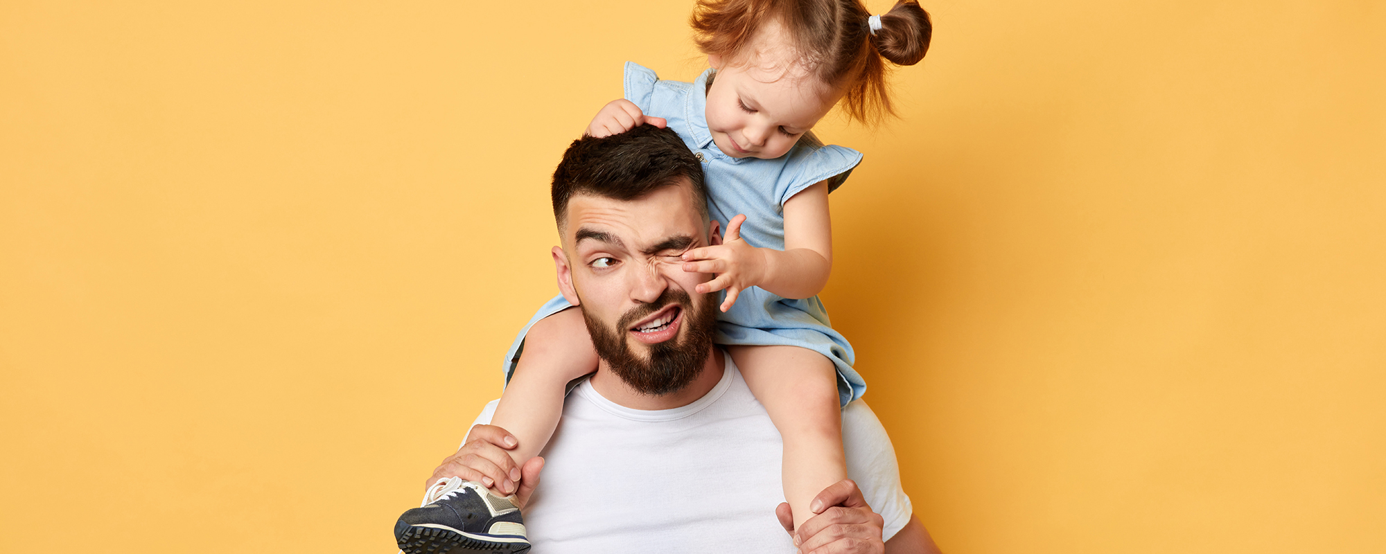 Papa hält kleine Tochter auf den Schultern. Tochter fährt mit Finger in dessen Auge.