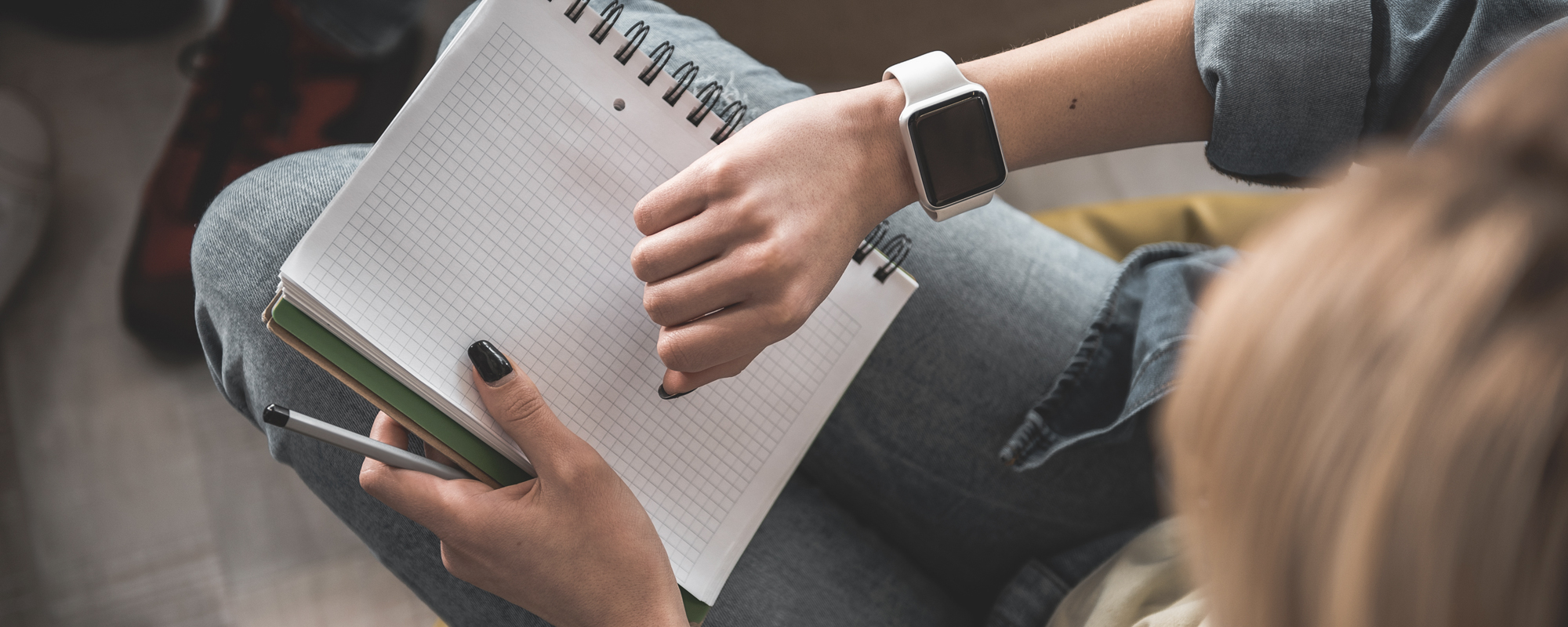 junge Frau mit leerem Notizblock auf dem Schoß blickt auf Smartwatch