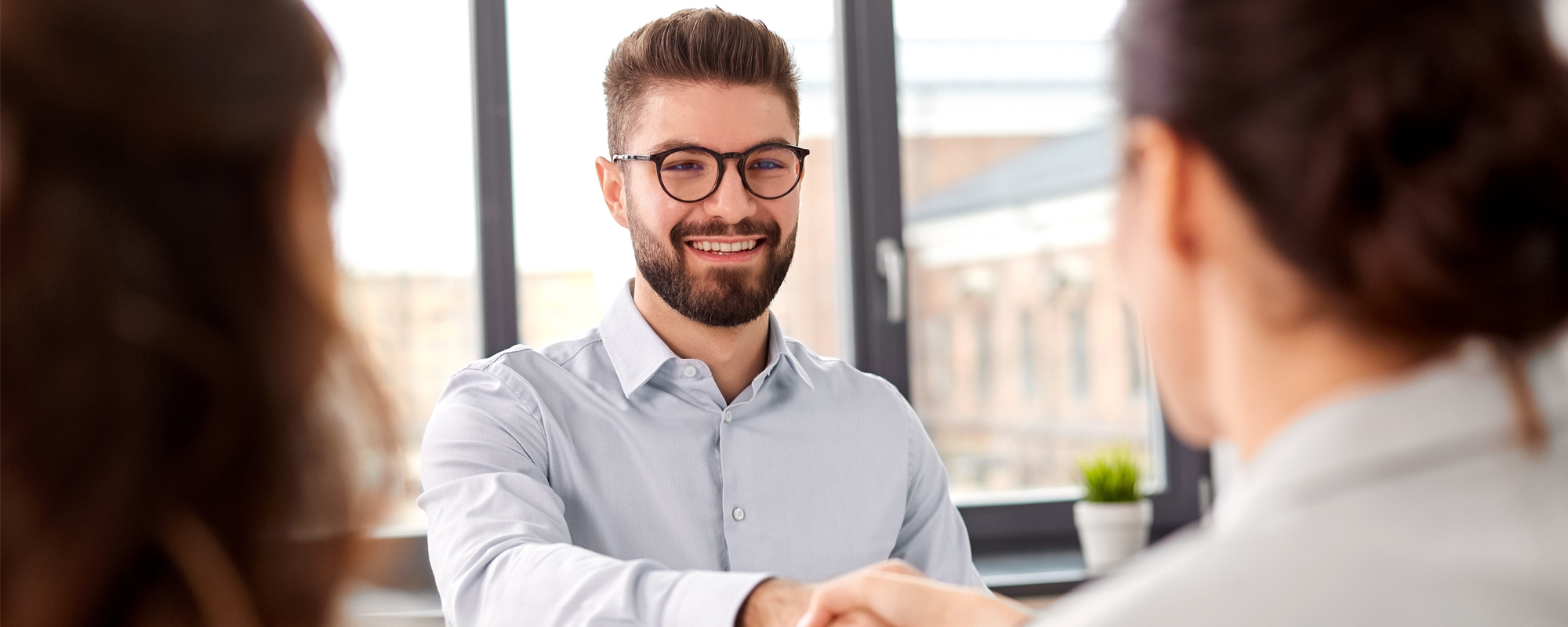 Zurück in der Arbeitswelt: Freundlich lächelnder Mann mit Brille und Hemd schüttelt zwei Frauen die Hand, die mit dem Rücken zur Kamera gewandt stehen.