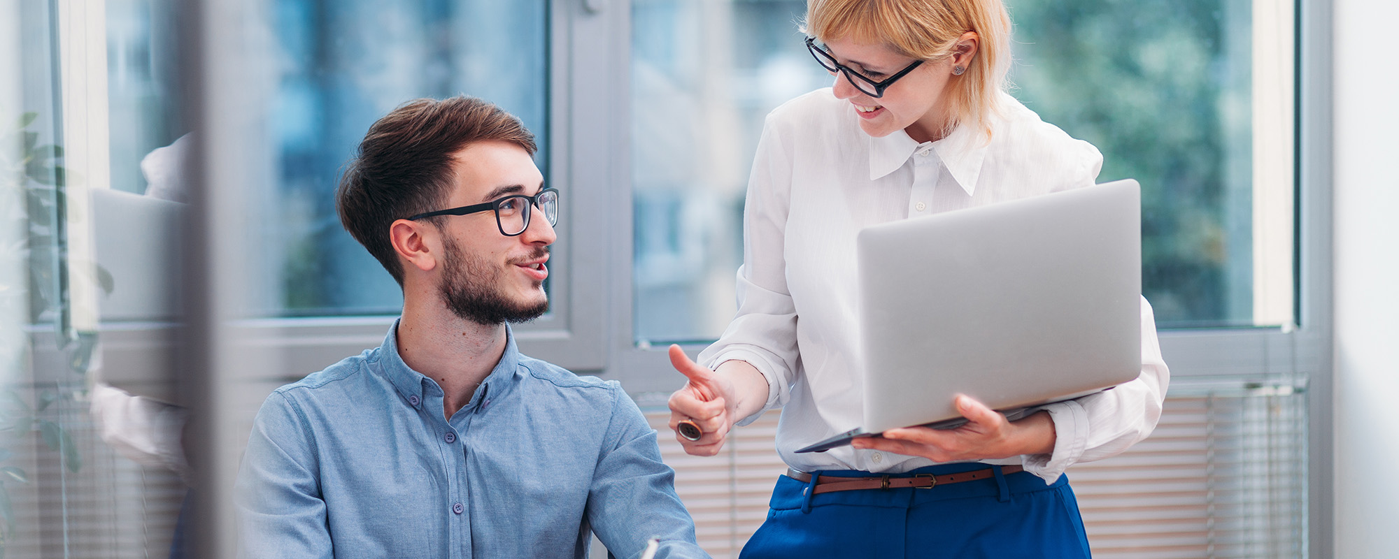 Blonde Frau im Businessoutfit steht im Büro neben Praktikanten, der am Schreibtisch sitzt, und zeigt lächelnd mit dem Daumen nach oben.