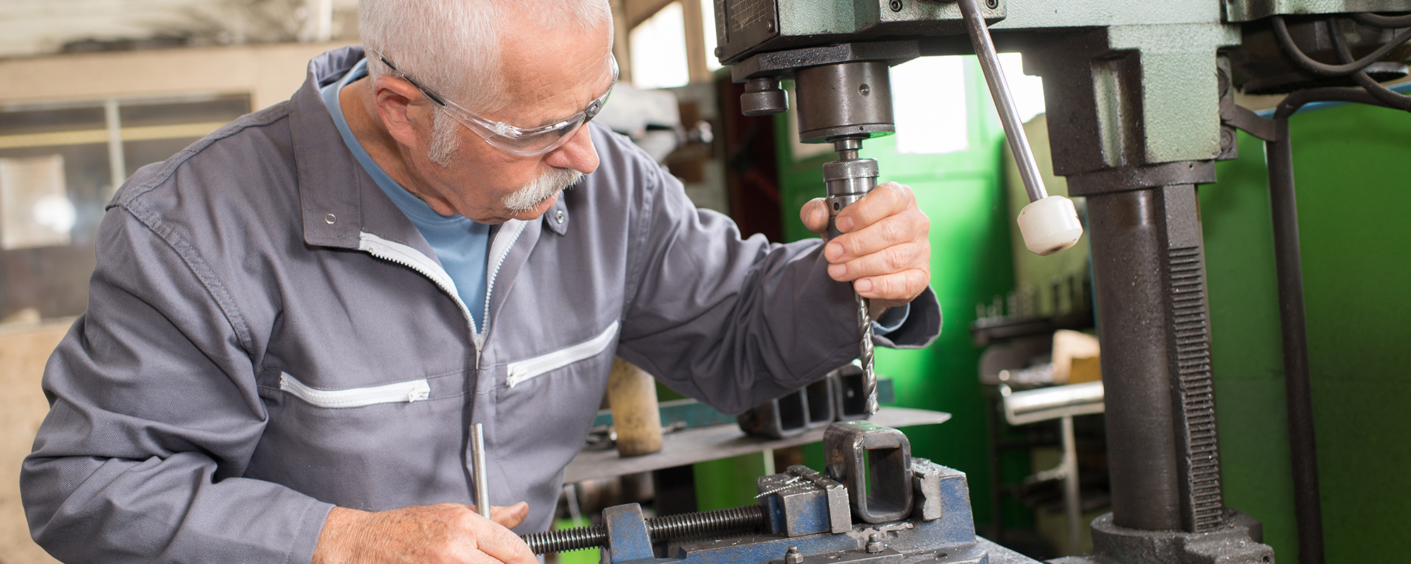 älterer Mann mit weißem schütteren Haar und Brille in Arbeitsmantel arbeitet an großer Maschine zur Metallverarbeitung.