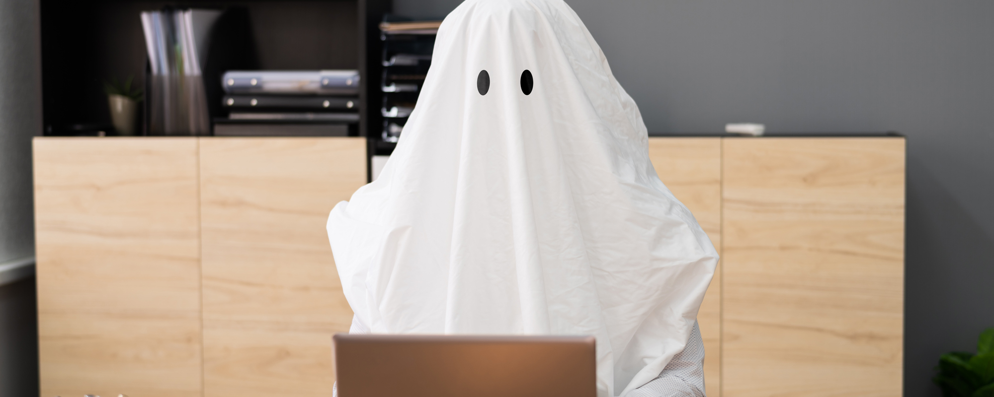 Job-Ghosting: Person in weißem Geist-Kostüm sitzt in einem Büro vor einem Laptop.