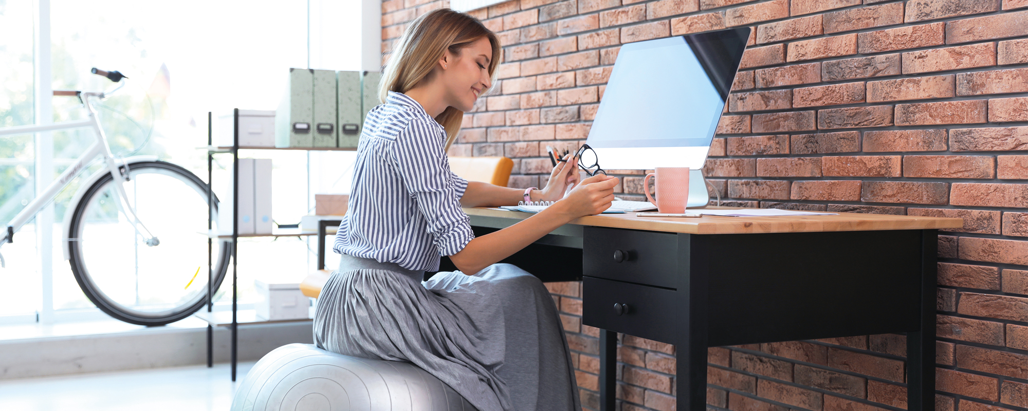 Junge Frau sitzt auf einem silbernen Sitzball in einem lichtdurchfluteten Büro lächelnd am Schreibtisch und arbeitet.