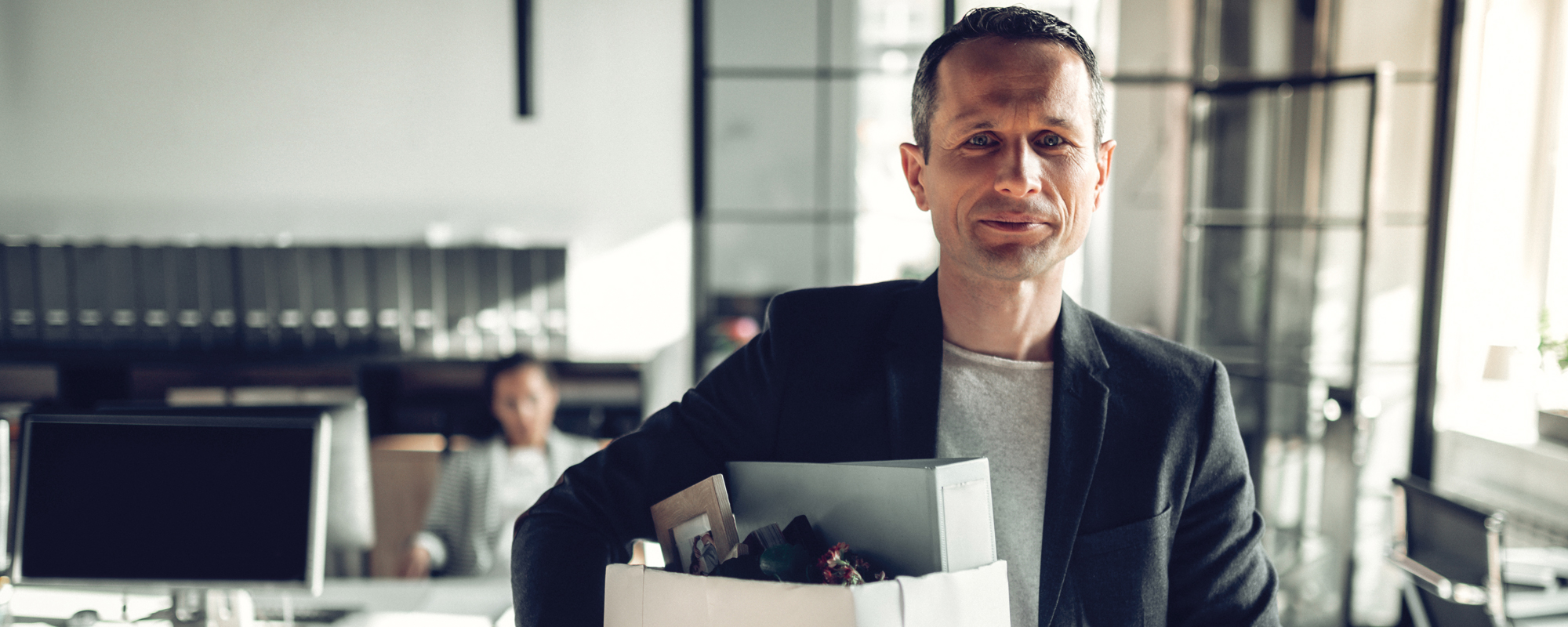 Offboarding: Mann in schwarzem Businessoutfit mit Karton unterm Arm verlässt das Büro.