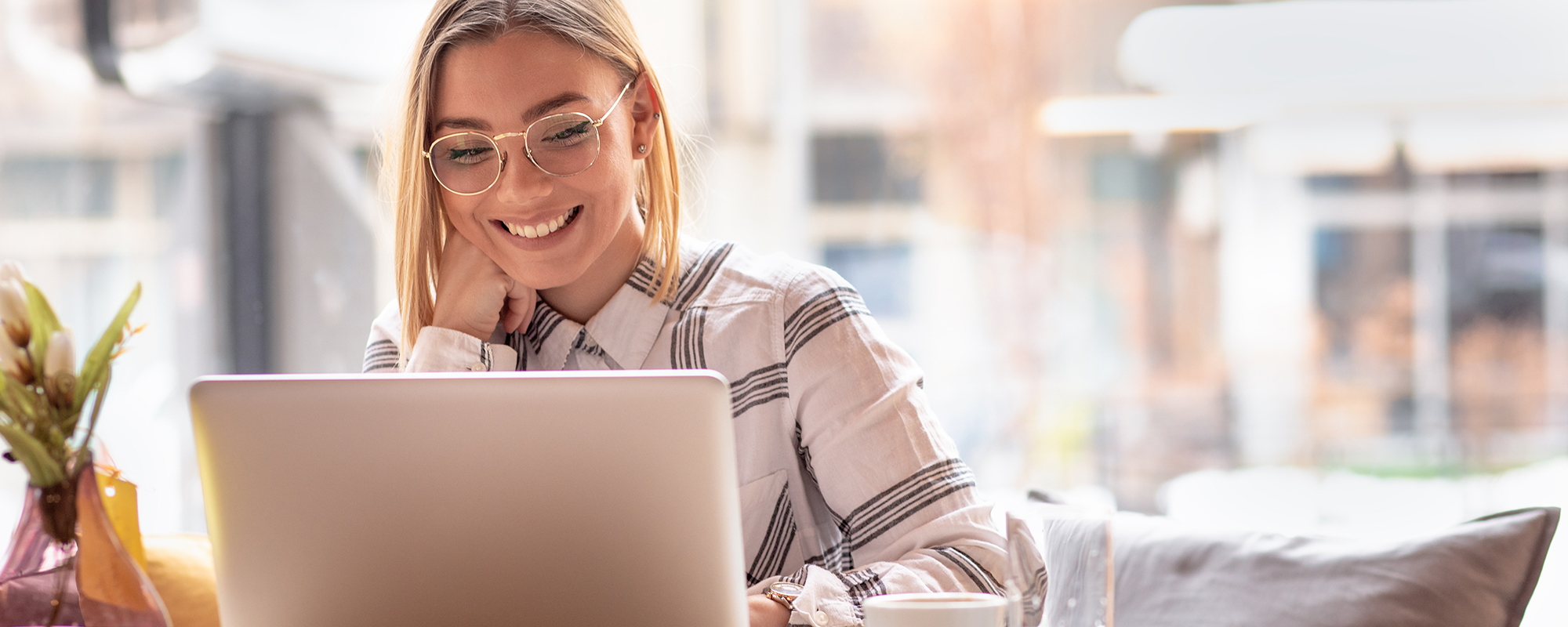 Junge blonde Frau mit Brille und weiß-kariertem Hemd sitzt mit ihrem Laptop vor dem Fenster in einem lichtdurchfluteten Café und arbeitet.