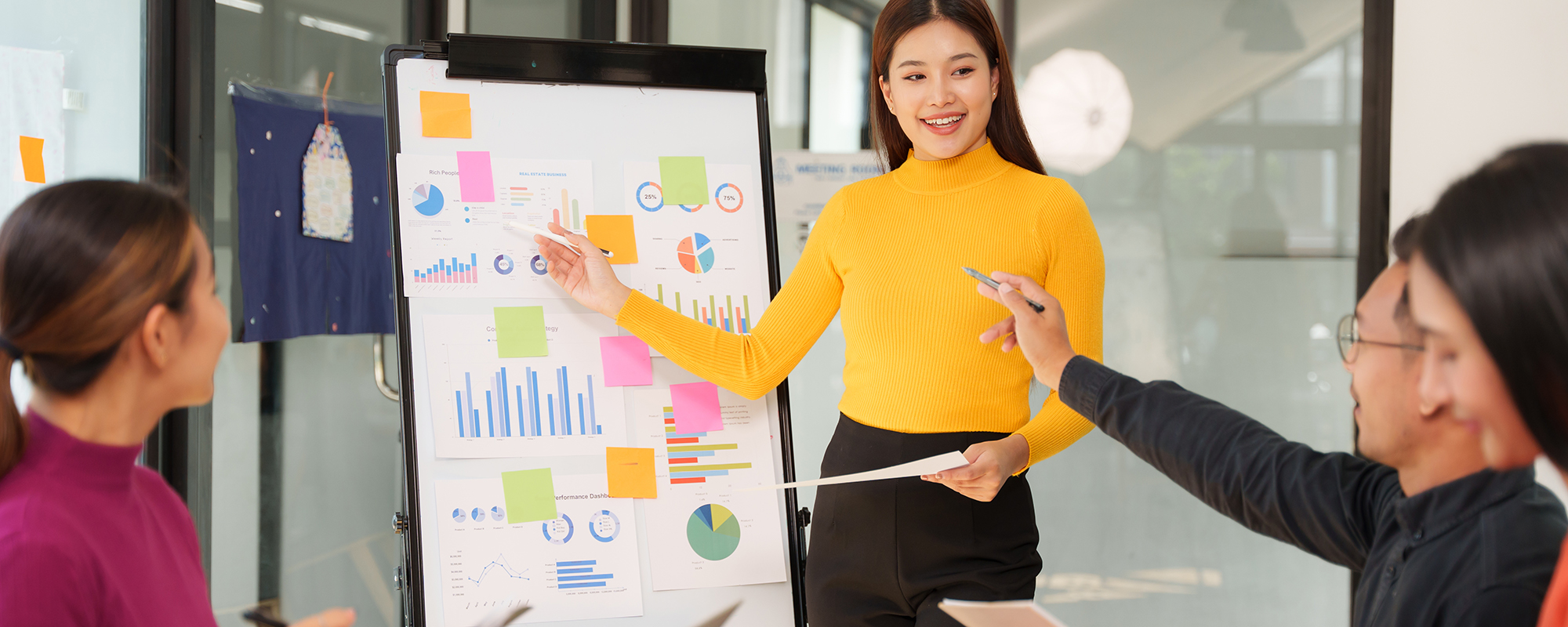 Coaching im Beruf: Asiatische Frau in gelbem Rollkragenpulli und Businessrock steht lächelnd an einem bunten Flipchart und erklärt den Anwesenden etwas.