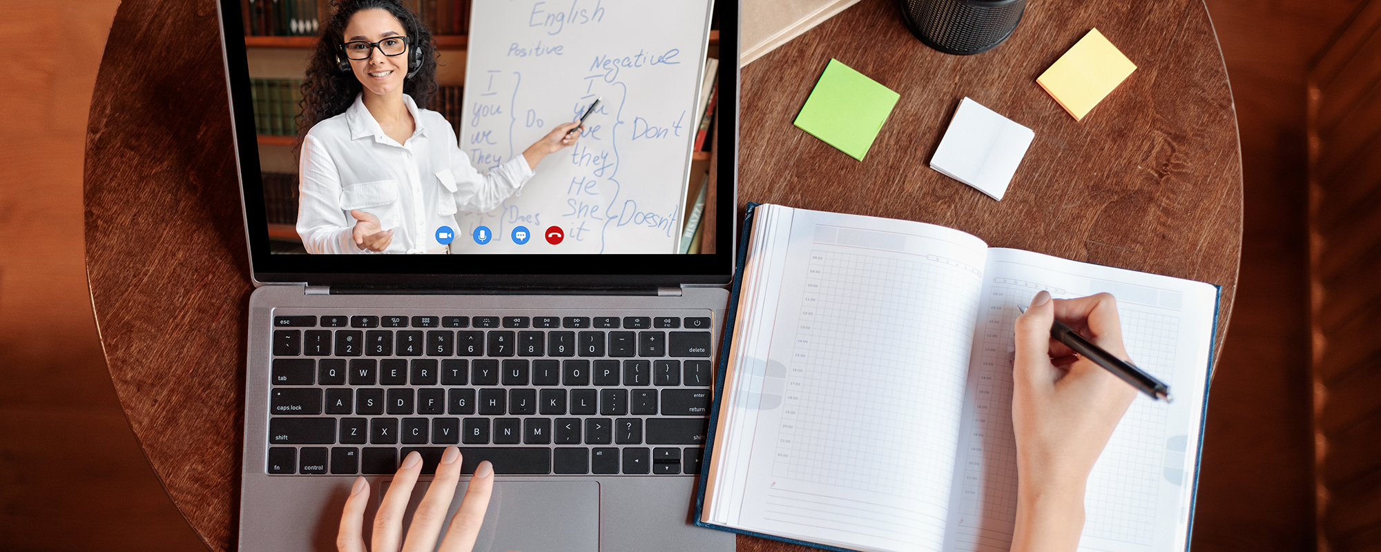 Person sitzt an einem runden Tisch und macht sich Notizen in ein Notizbuch, während auf dem Laptop ein Online-Kurs läuft.