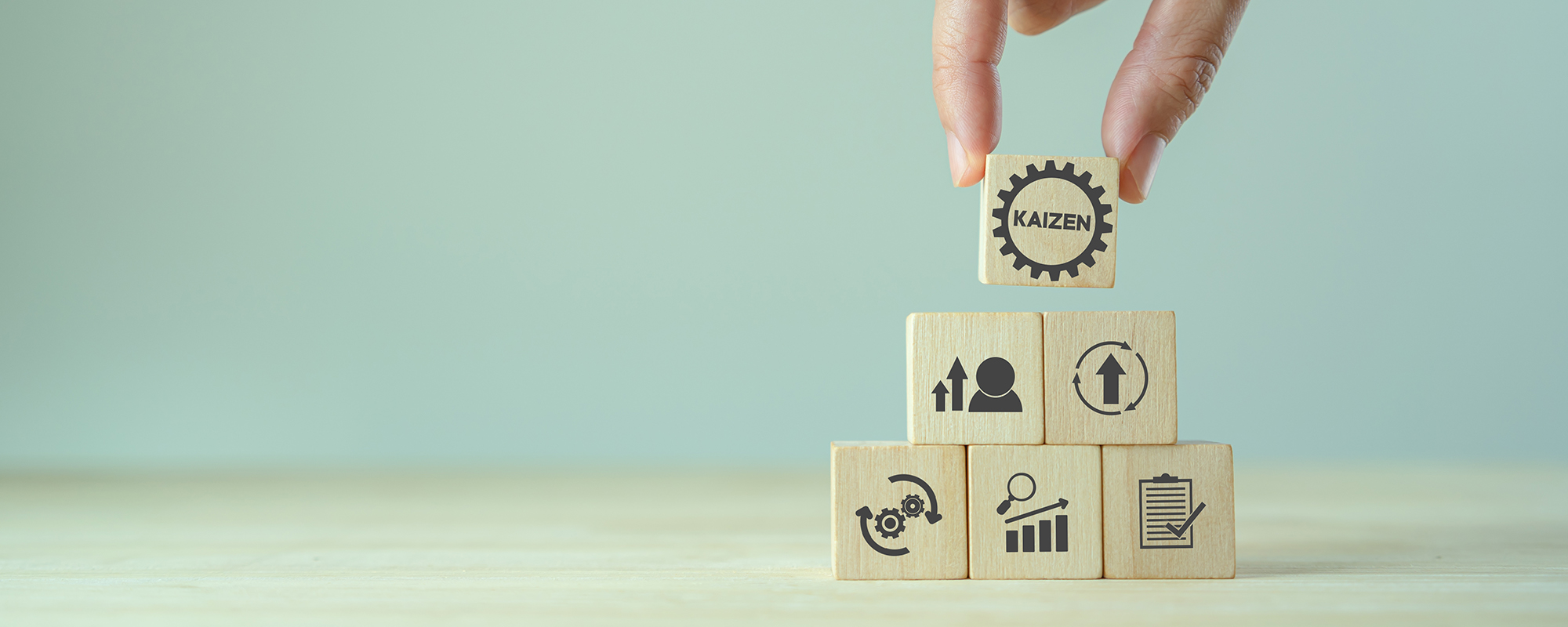 Ein Turm aus Würfeln mit Symbolen, die die fünf Säulen des Kaizen-Prinzips symbolisieren. Eine Hand legt einen sechsten Würfel mit dem Wort „Kaizen“ ganz oben auf den Stapel.