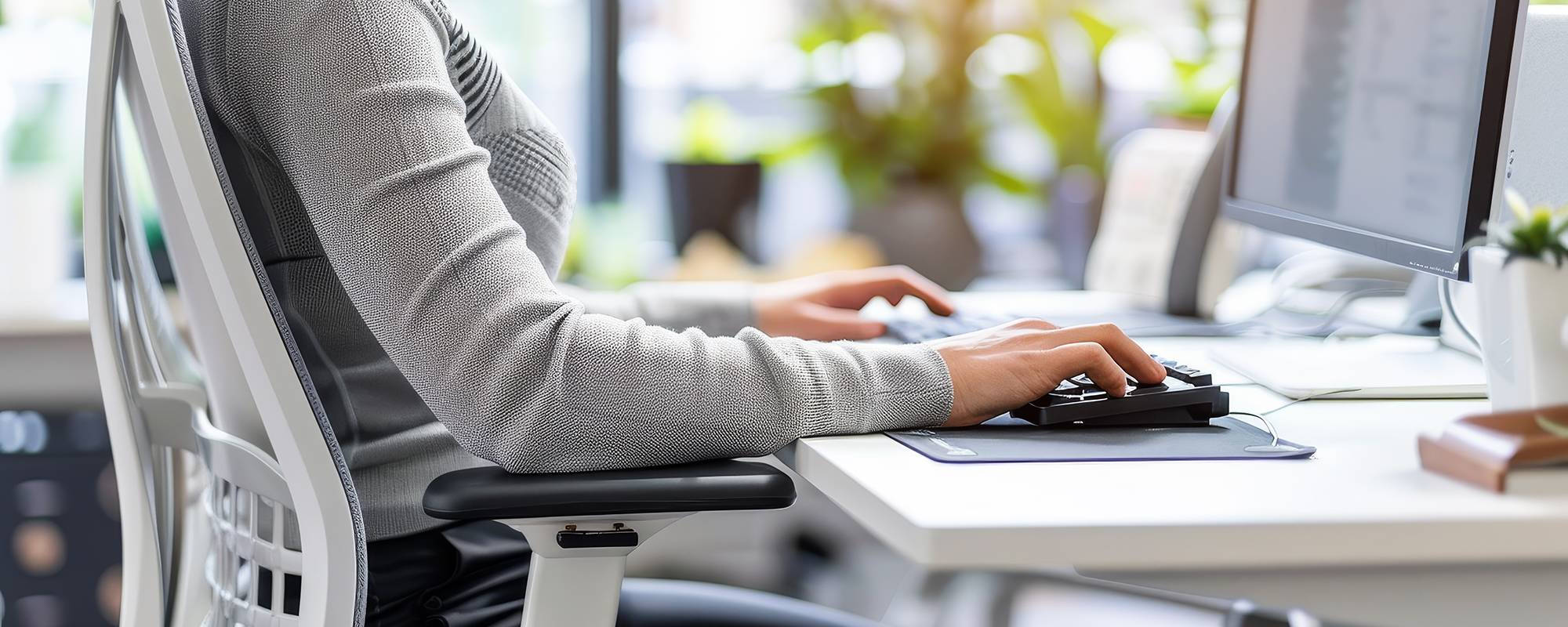 Frau sitzt in einem lichtdurchfluteten Büro auf einem ergonomischen Bürosessel und arbeitet am Schreibtisch auf ihrem Computer.