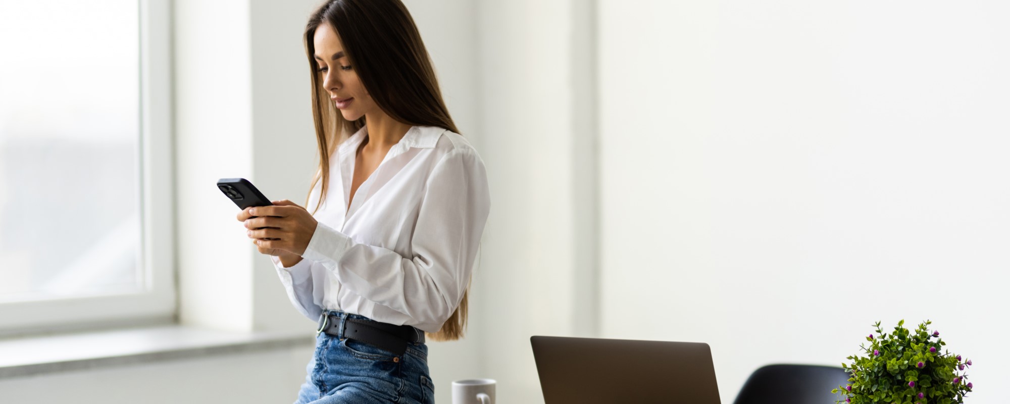 Junge Frau mit Smartphone in der Hand steht entspannt vor einem Fenster, neben einem Laptop und einer Pflanze auf einem Arbeitsplatz. Symbolbild zur Handynutzung am Arbeitsplatz, thematisch passend zu Regelungen, Erlaubtem und Grenzen im beruflichen Kontext.