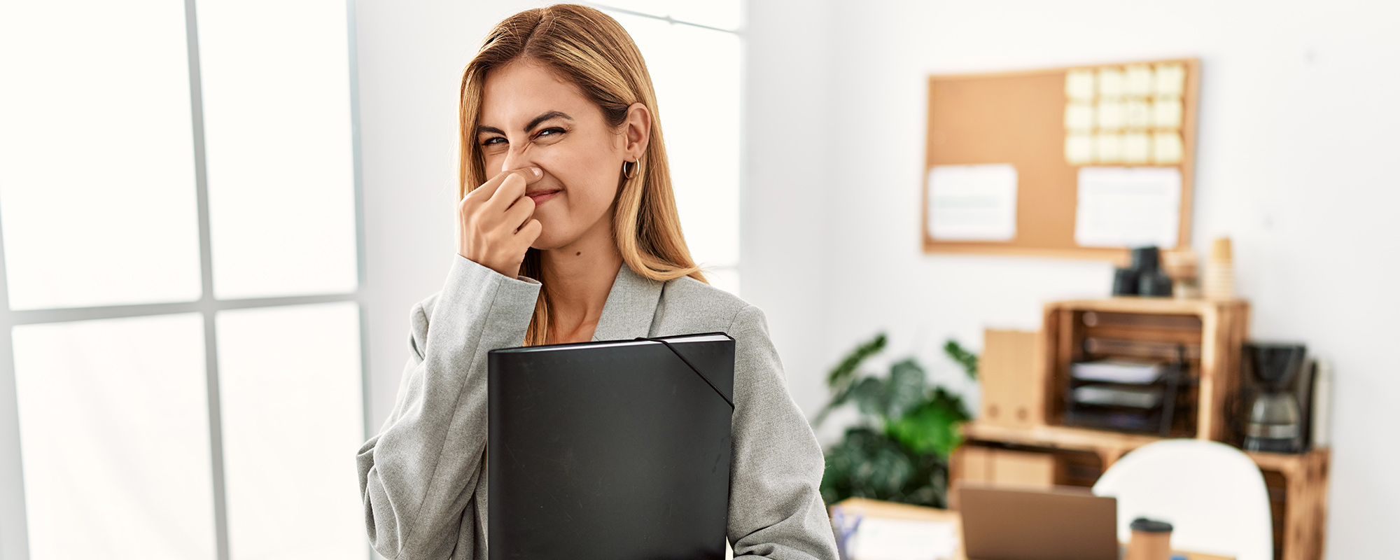 Blonde Geschäftsfrau im Büro hält sich wegen unangenehmem Geruch in der Büroküche die Nase zu. Etikette in der Büroküche und mangelnde Hygiene in Gemeinschaftsräumen.