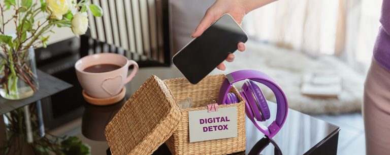 Frau legt ihr Smartphone in Korb mit Technikgeräten auf Tisch. Digital Detox im Büro und Zuhause, Auszeit von Handy und Bildschirm, für weniger Technologieabhängigkeit.