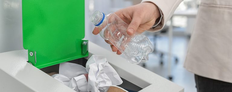Frau wirft eine benutzte Wasserflasche in einen vollen Mülleimer mit Papiermüll in einem Office ohne Zero-Waste-Kultur.