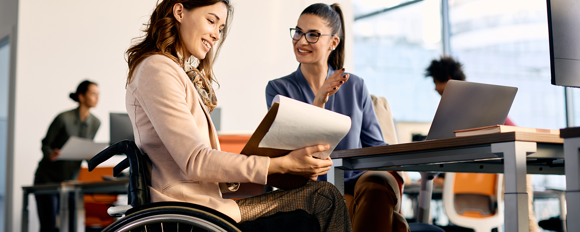Eine Bewerberin im Rollstuhl im positiven Bewerbungsgespräch mit einer Personalerin, was die rechtliche Frage aufwirft, ob Fragen zur Krankheit im Vorstellungsgespräch zulässig sind.