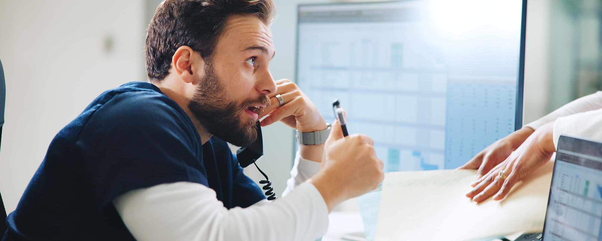 Ein Mann beim Multitasking im Büroalltag. Er telefoniert, arbeitet am Computer und interagiert mit Kollegen, ein Symbol für Produktivität und Teamarbeit.