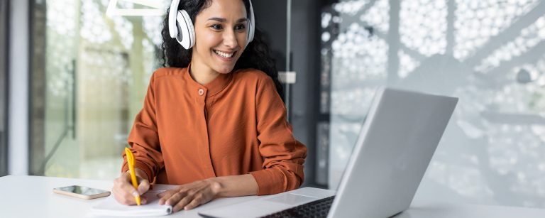 Frau mit Kopfhörern sitzt an einem Schreibtisch und macht sich begeistert Notizen bei Ihrem Online-Kurs zur beruflichen Weiterbildung.