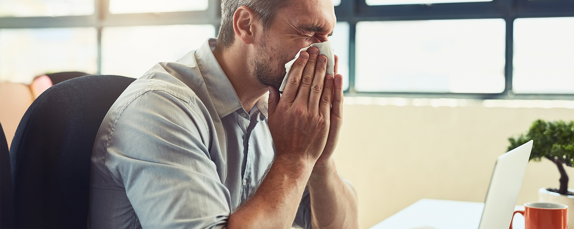 Mann sitzt schneuzend vor dem PC im Büro. Krank arbeiten ist keine gute Idee.