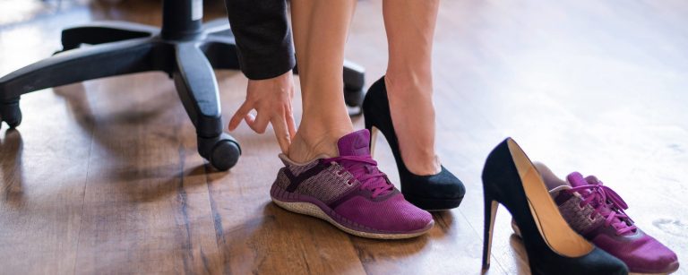 Frau in einem Büro wechselt von ihren professionellen Pumps in violette Sportschuhe für einen revitalisierenden Spaziergang in der Mittagspause.