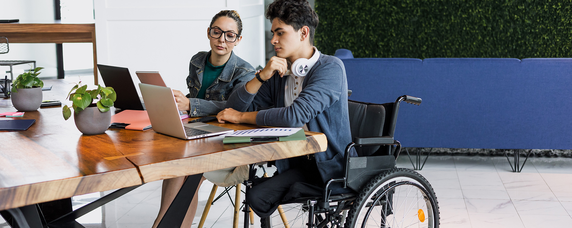 Junger Mann in Rollstuhl sitzt mit Frau ohne Behinderung an Schreibtisch und arbeitet.