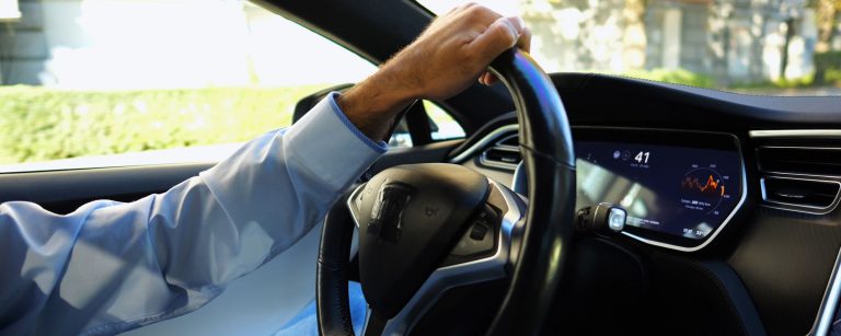 Ein Pendler in blauem Hemd sitzt auf dem Weg zur Arbeit in seinem Elektroauto und hat eine Hand locker auf das Lenkrad gelegt, während draußen die Sonne scheint.